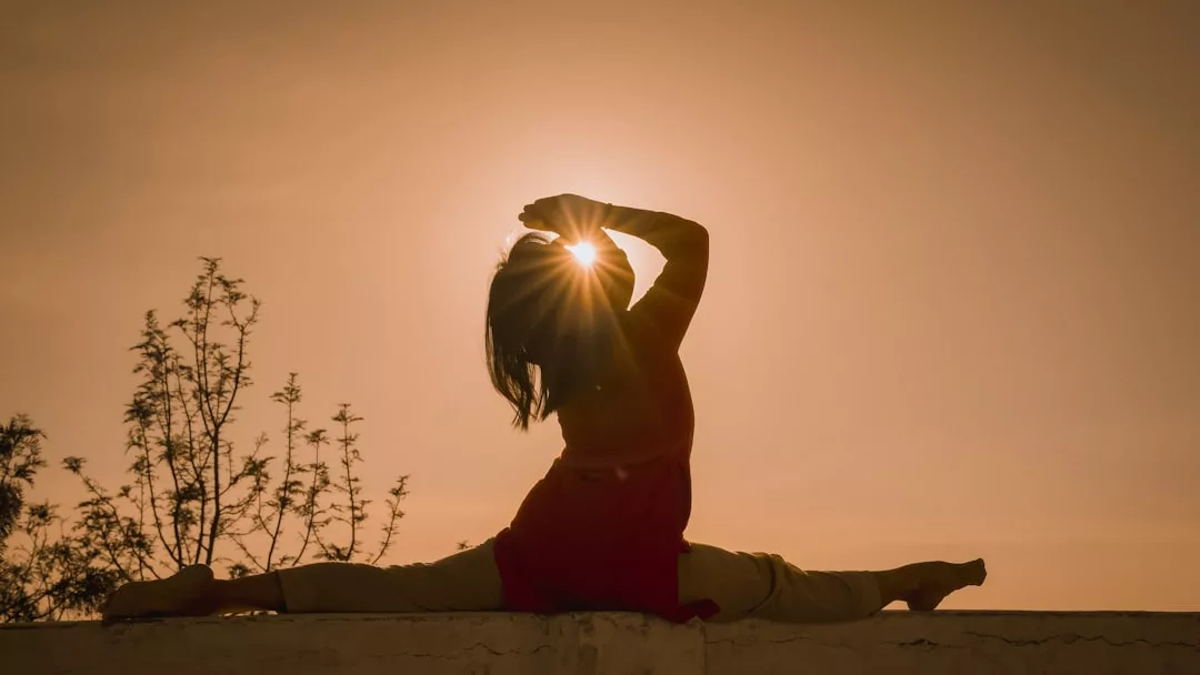 Person performing a split yoga pose on a ledge at sunset in Majorca, with the sun positioned behind their head and trees in the background, capturing a sense of luxury lifestyle amidst breathtaking scenery.