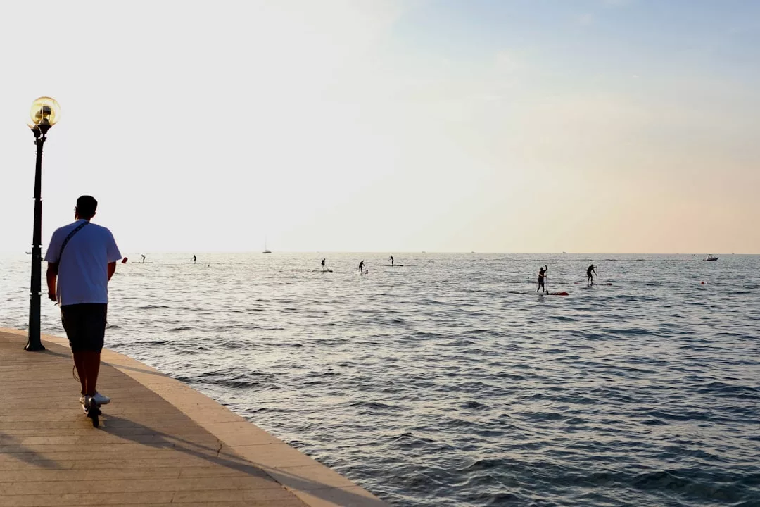 A person strolls along a seaside promenade in Majorca, while several people paddleboard on the calm water under a clear sky, enjoying the island’s luxury lifestyle.