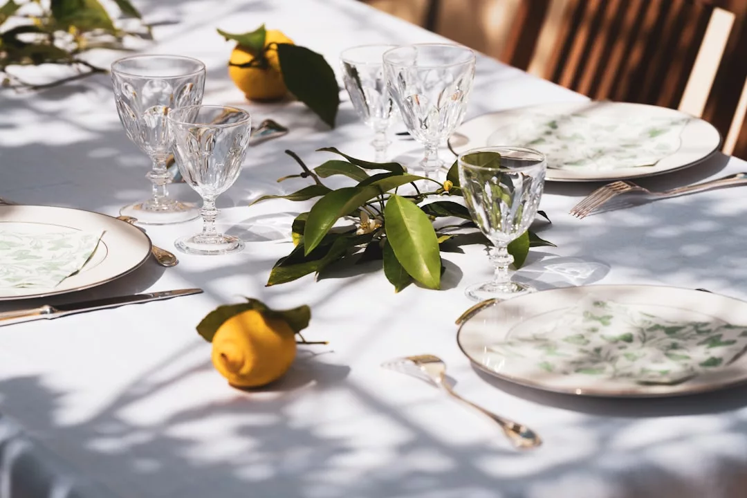 A table set with patterned plates, glasses, forks, and knives, decorated with lemons and green leaves on a white tablecloth—perfect for enjoying a touch of Majorca luxury and lifestyle.
