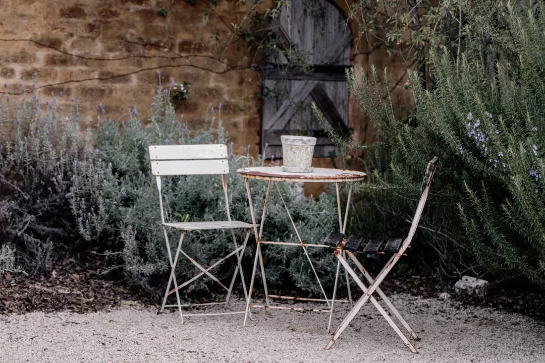 Two metal chairs and a small round table with a potted plant are set on a gravel area, surrounded by green bushes and a weathered stone wall with a wooden door, evoking the relaxed luxury lifestyle of Majorca.
