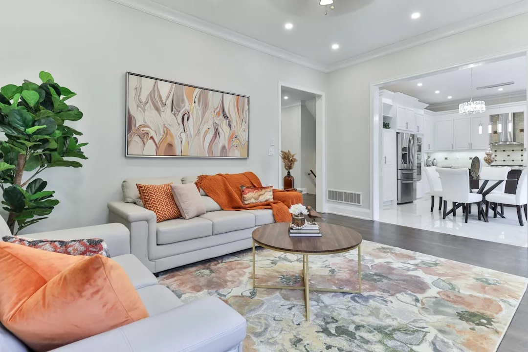 Experience luxury living in this modern Majorca-inspired living room, featuring a white sofa, orange accents, round coffee table, abstract wall art, and seamless flow into a stylish kitchen and dining area.
