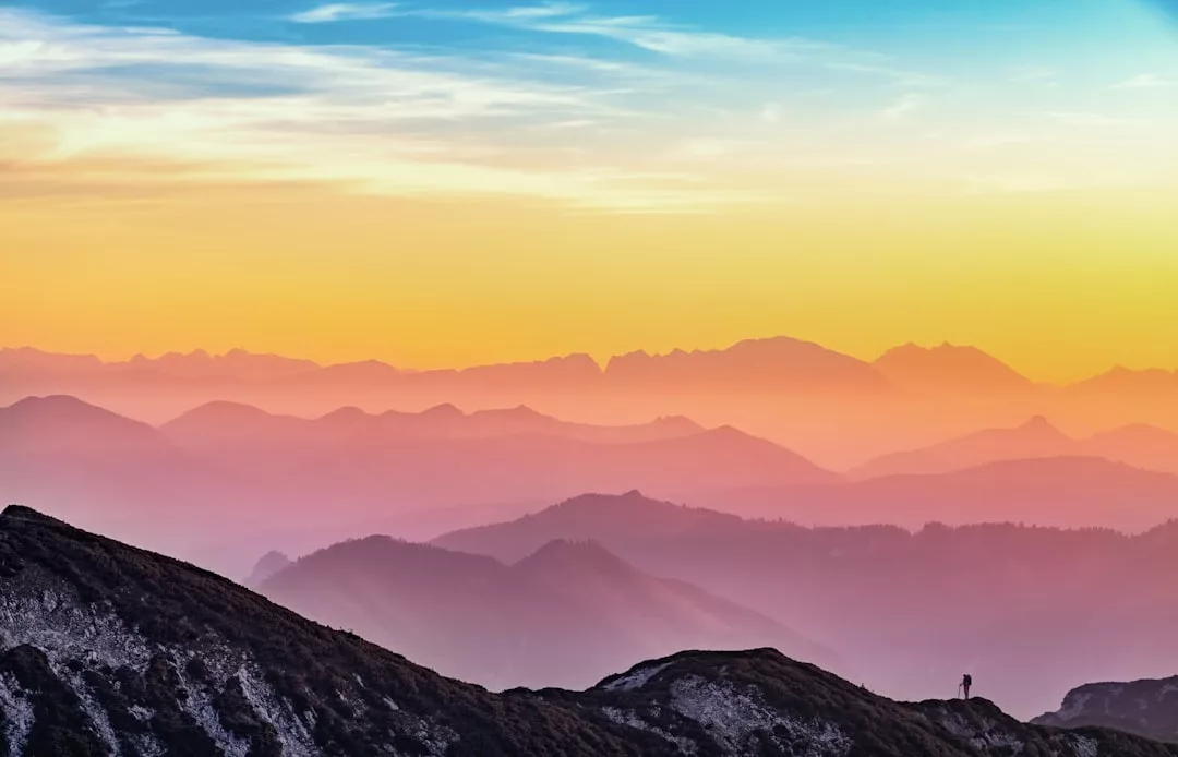 A person stands on a mountain ridge in Majorca, overlooking layered hills and distant peaks under a colorful sunset sky, capturing the essence of luxury lifestyle amid blue, yellow, and orange hues.