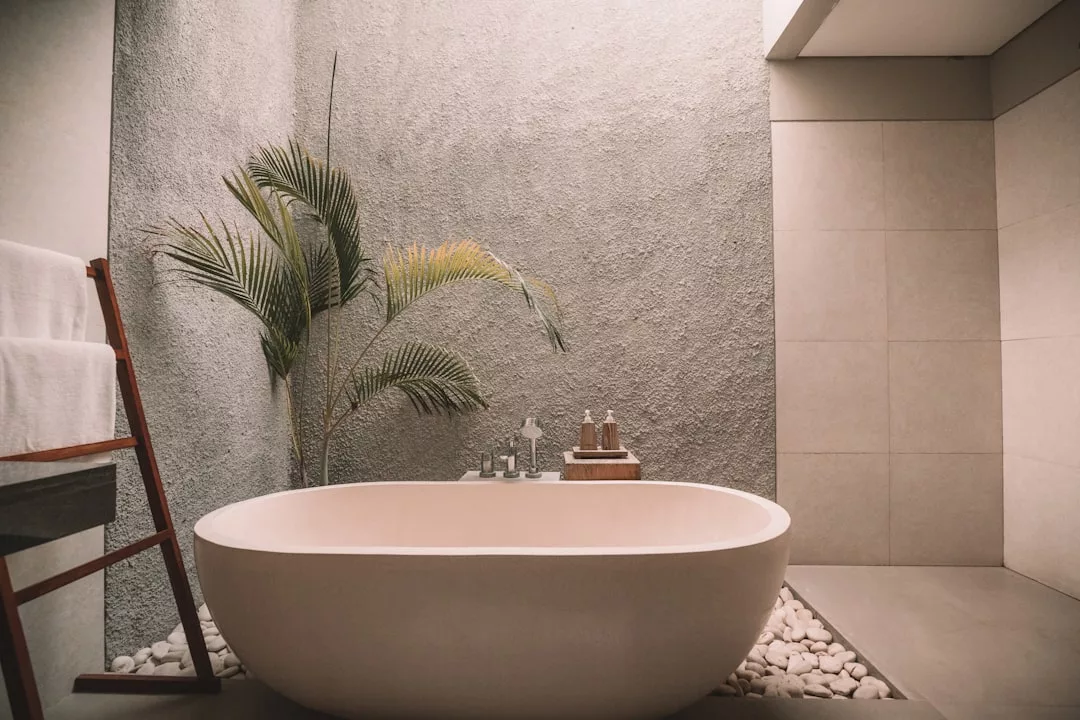 Modern bathroom with a freestanding bathtub on white pebbles, evoking a sense of luxury. A plant rests against the textured wall, while a towel rack on the left completes this serene lifestyle-inspired space.