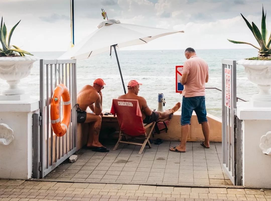 Three men, two seated under a beach umbrella and one standing, gather by a gated seaside area in Majorca with ocean views. Nearby, luxury touches like potted plants and lifesaving equipment highlight an elegant coastal lifestyle.