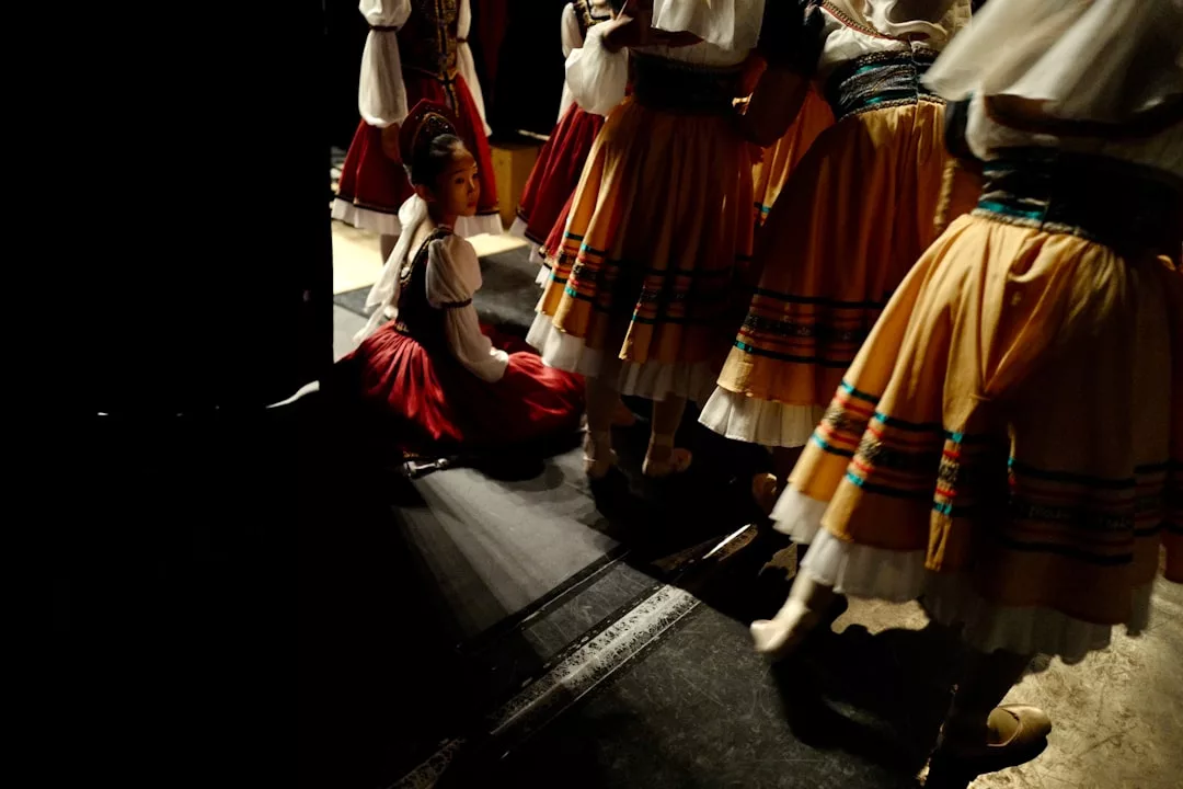 A girl in traditional dress kneels backstage while other performers in similar costumes walk past her under stage lighting, capturing a glimpse of the vibrant lifestyle and cultural richness reminiscent of Majorca.