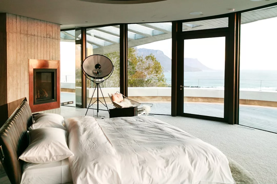 A modern bedroom with a double bed, large windows, a fireplace, lounge chair, and a stunning ocean view with mountains in the background—perfect for embracing a luxury lifestyle inspired by Majorca.