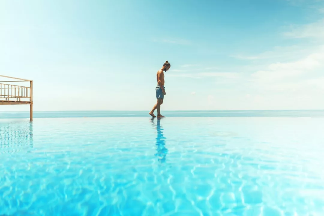 A man in swim shorts stands at the edge of an infinity pool, looking down, with a clear sky and calm sea in the background—capturing the essence of luxury lifestyle in Majorca.