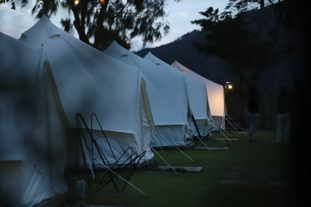 A row of white canvas tents set up on grass at dusk evokes a sense of luxury lifestyle, with a few standing figures visible in the background near trees and a hillside.