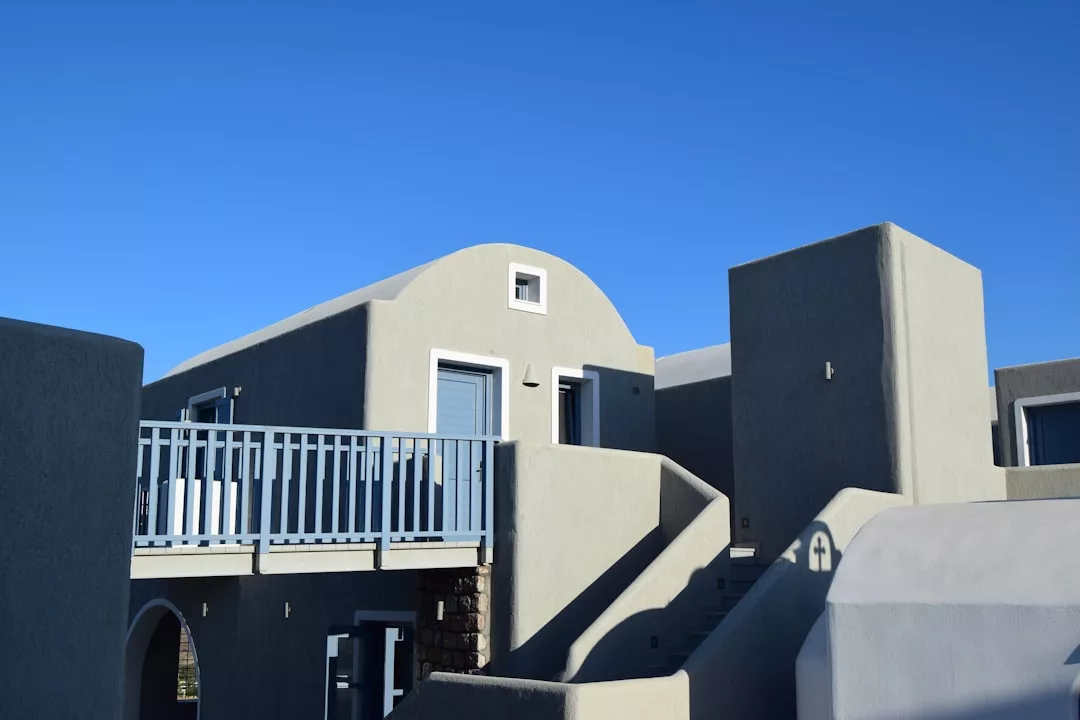 A modern, light gray stucco building with blue doors, windows, and a balcony railing under a clear blue sky evokes a luxury lifestyle inspired by the elegance of Majorca.