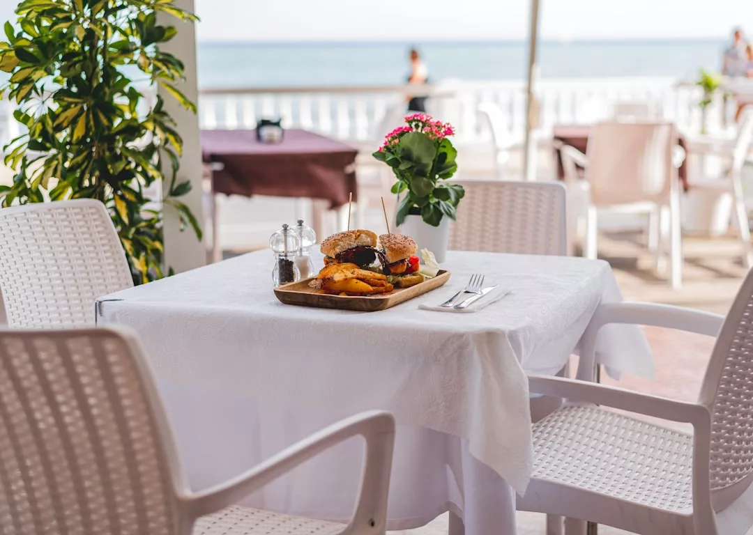 A table with a white tablecloth set for one holds a meal of burgers and fries, a fork and knife, condiments, and a small flowerpot, capturing the relaxed lifestyle of Majorca with an ocean view in the background.