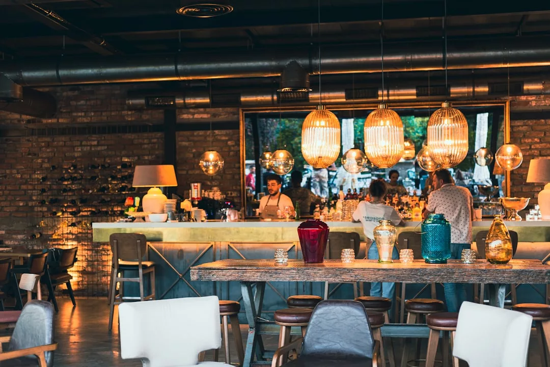 Modern bar interior with pendant lights, wooden tables, and assorted chairs evokes a luxury lifestyle vibe. People stand behind the counter, while brick walls and large windows in the background add to the chic Majorca-inspired ambiance.