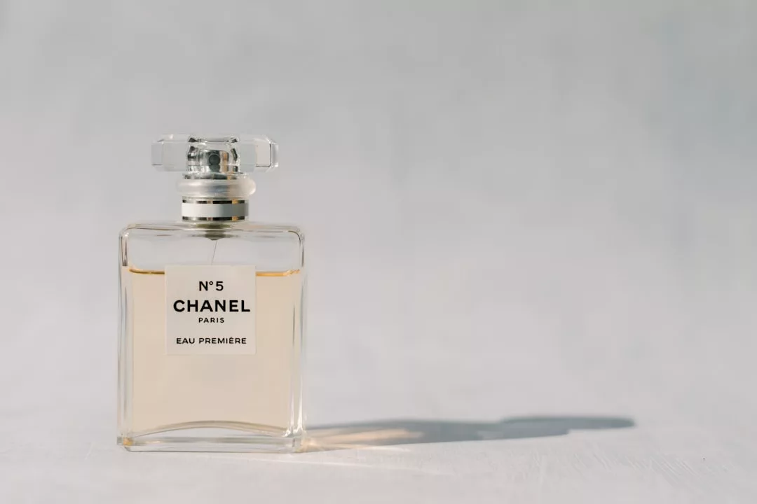 A clear glass bottle of Chanel No. 5 Eau Première perfume sits on a light surface with a shadow cast to the right, evoking a sense of luxury and effortless lifestyle inspired by sunlit days in Majorca.