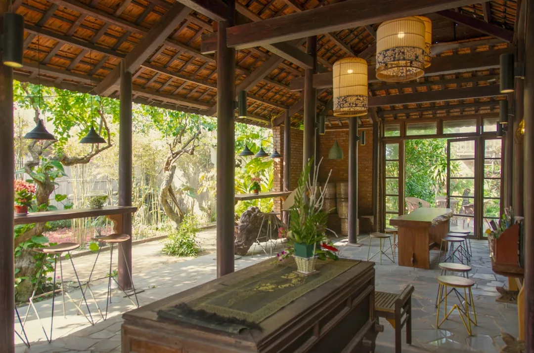 Open-air room with wooden beams, tiled roof, and large windows; features plants, pendant lights, and wooden tables overlooking garden views—a touch of luxury that captures the relaxed lifestyle of Majorca.