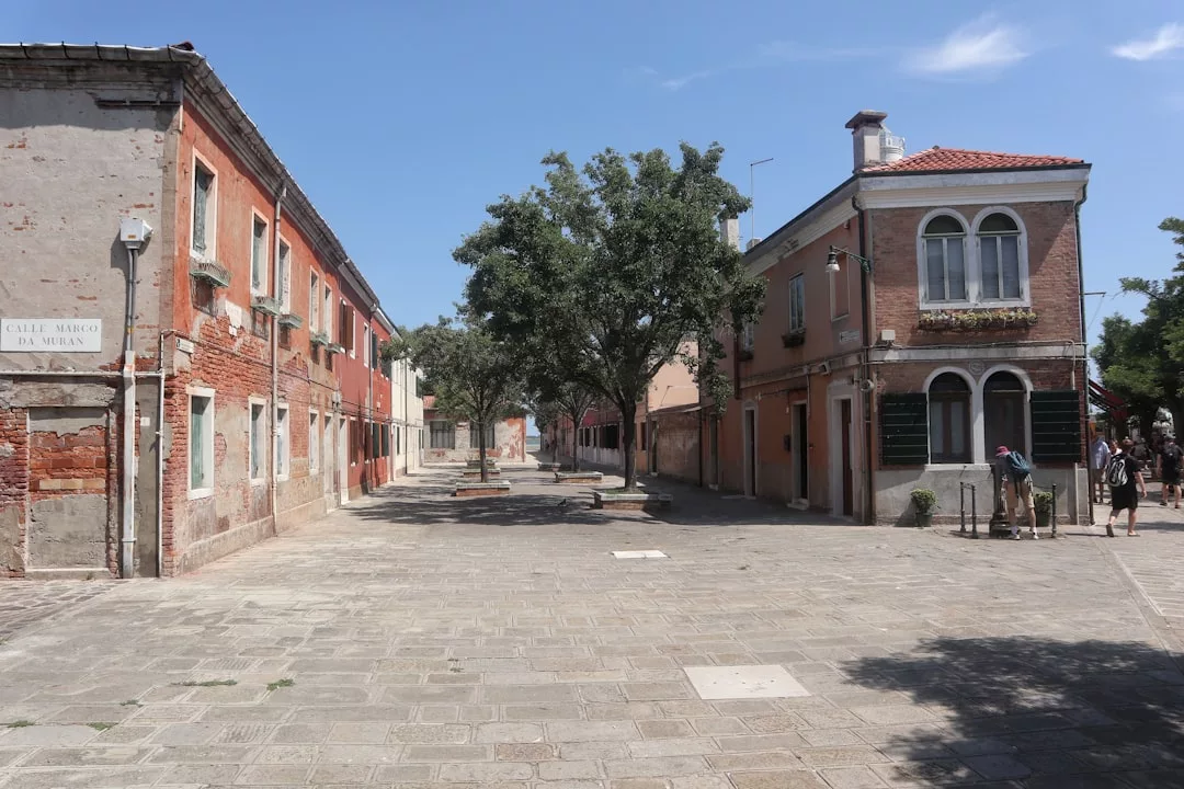 A quiet, sunlit street in Majorca lined with old brick buildings and a large tree in the center, exuding a sense of luxury lifestyle as a few people stroll in the distance.