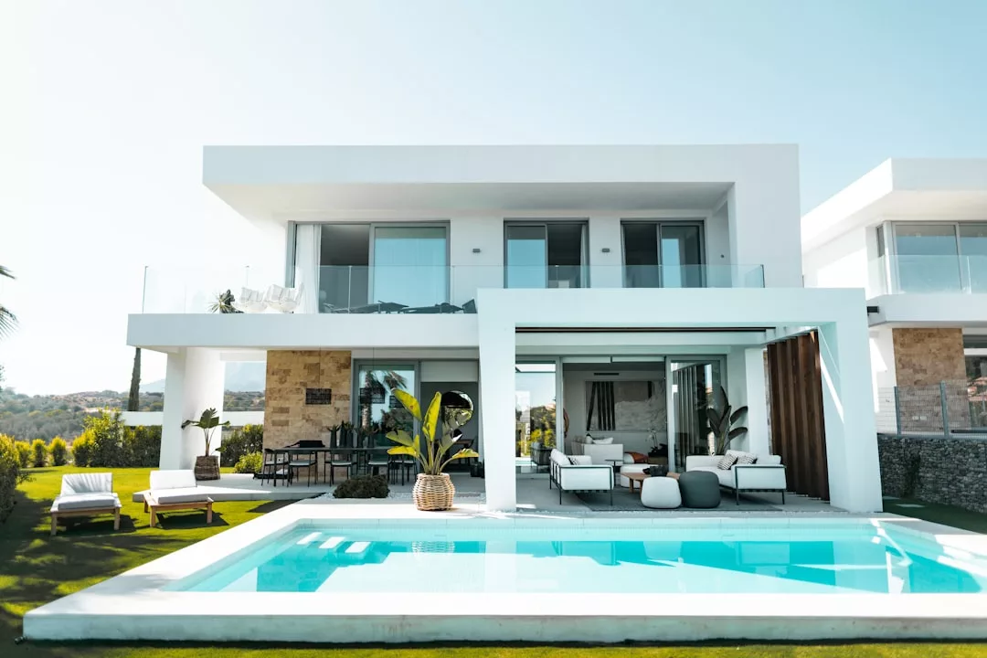 Modern two-story house with large windows, a covered patio, lounge chairs, and a swimming pool in the backyard—perfect for indulging in a luxury Majorca lifestyle on a sunny day.