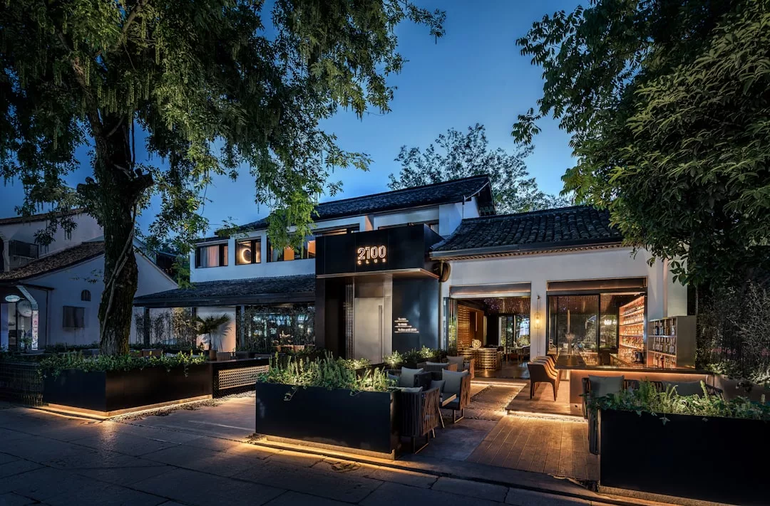 Modern restaurant exterior at dusk with large windows, outdoor seating, greenery, and warm lighting offers a taste of luxury lifestyle, surrounded by tall trees and a paved walkway inspired by the elegance of Majorca.