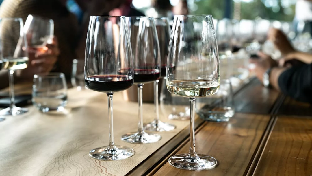 A row of wine glasses filled with red and white wine sits on a wooden table, capturing the essence of luxury lifestyle, with people in the background out of focus.
