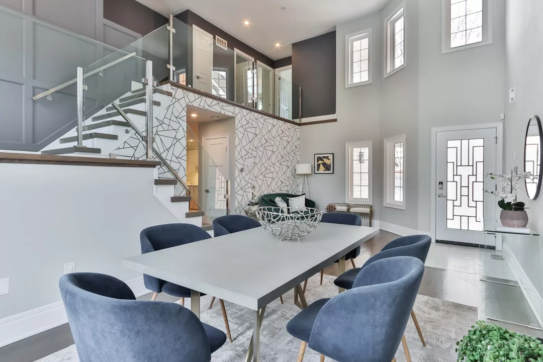 Modern dining room with a gray table, six blue velvet chairs, a decorative centerpiece, and a staircase with glass railing leading to an upper floor—this bright, open space exudes luxury and effortlessly elevates your lifestyle in Majorca.