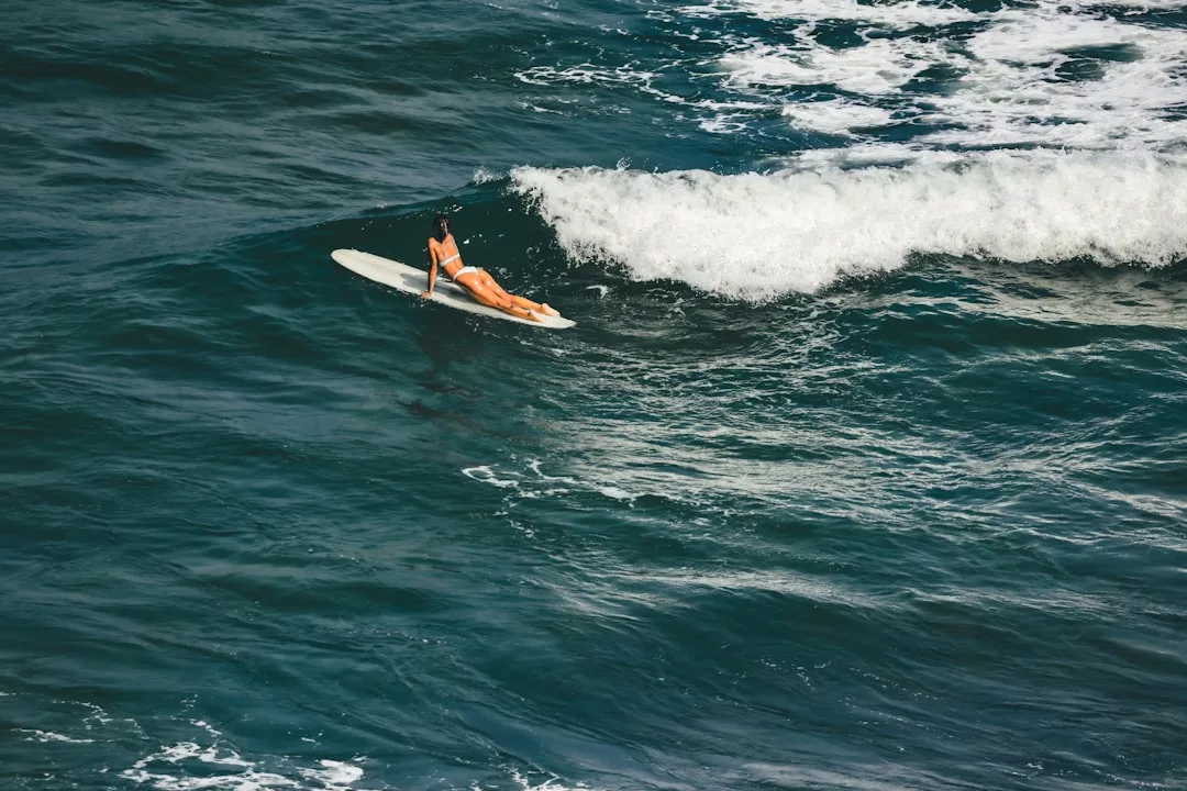 Embracing a luxury lifestyle, a person in an orange swimsuit lies on a surfboard in the tranquil ocean off Majorca, facing incoming waves.
