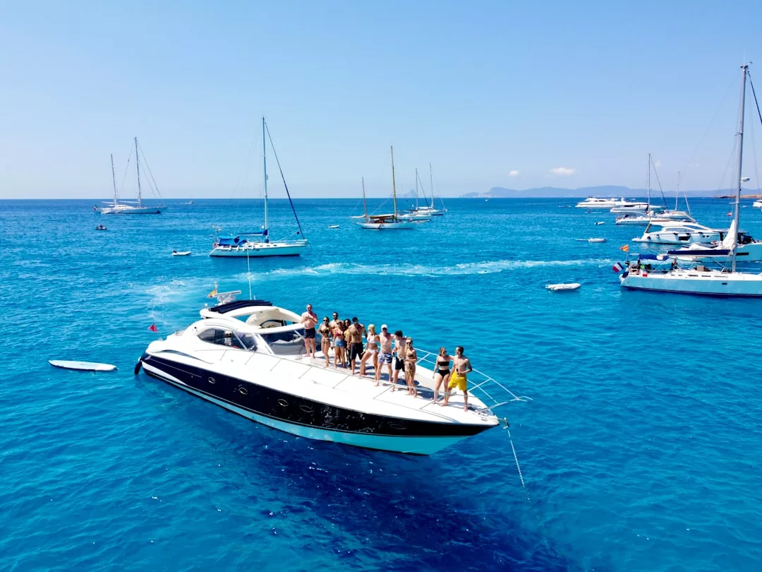 A group of people enjoy a luxury lifestyle on the deck of a white yacht in clear blue water near Majorca, surrounded by several other boats under a sunny sky.